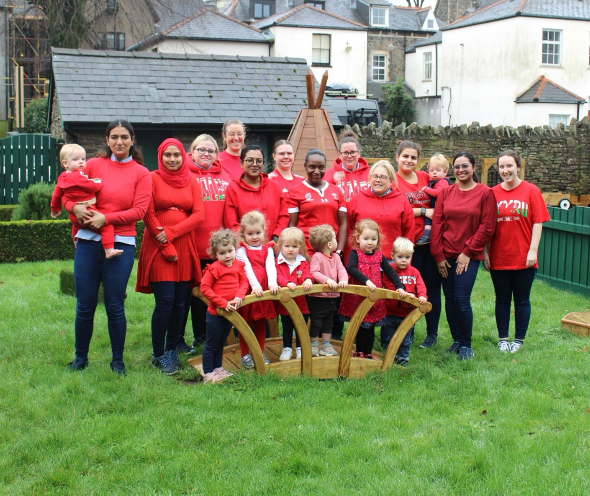 Acorns Nurseries A group of adults and children in red gathers on the grass near a wooden play structure, fostering community engagement against the backdrop of charming houses.